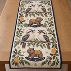 Four illustrated laughing kookaburras with broad bills and barred tails frame two brown toy rocking wombats with ochre saddles and navy rockers, orange halves, cedar, holly, mistletoe and ochre snowflakes on ivory with navy binding. Flat printed Christmas artwork. Includes one table runner. Quilted cotton hero with visible sewn quilting and fabric drape.